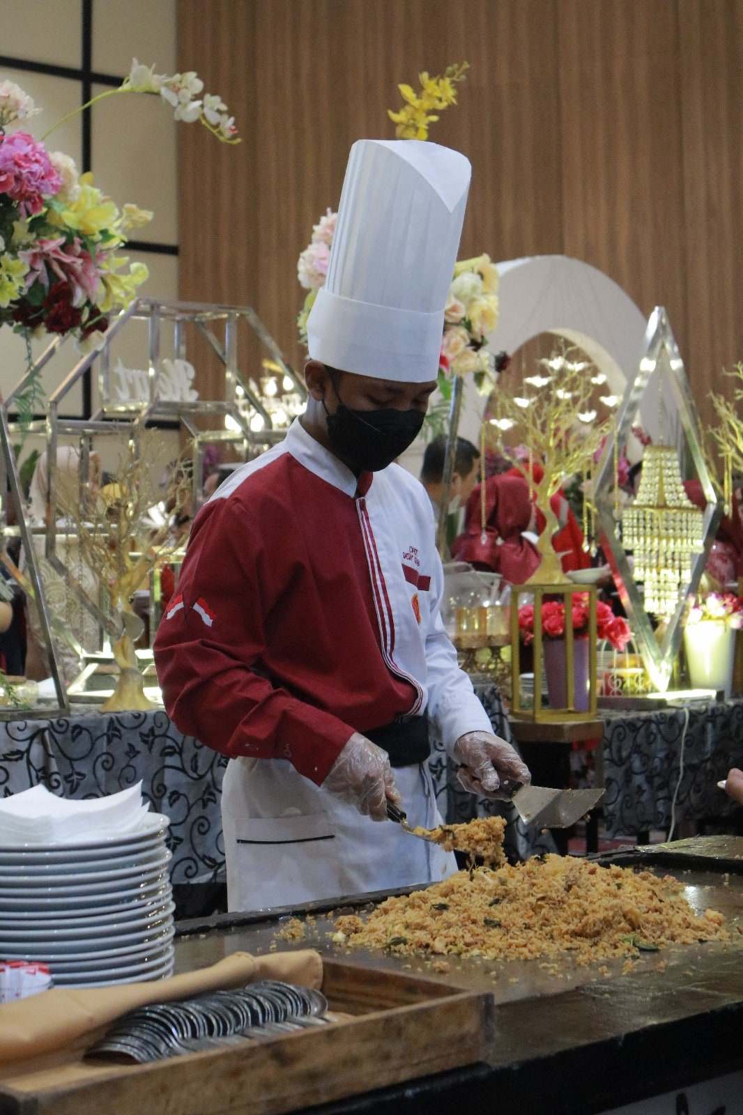 Koki bertopi tinggi mengaduk nasi goreng di atas wajan datar saat acara berlangsung